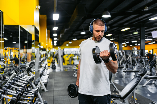 Handsome young muscular man training in the gym with dumbbells while listening to music via headphones in the Kuwait City.