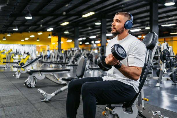 young man training with dumbbells while listening to music in the gym - weight training body building men human muscle - fotografias e filmes do acervo