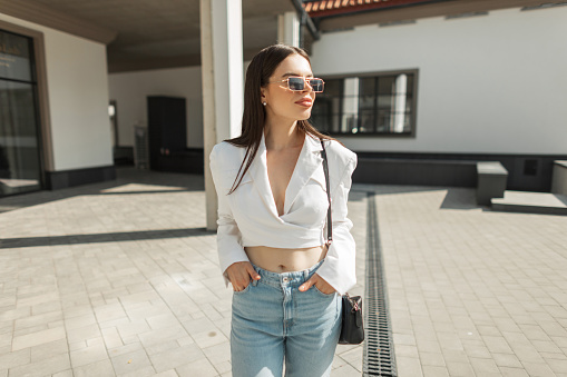 Cool fashionable American prett woman model with vintage sunglasses in trendy outfit with jacket and blue jeans with bad walks on the street on sunny day. Beautiful girl