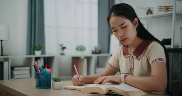 istock Young asian woman sitting at desk holding pen making notes in diary and finding information in books at home. 2014797712