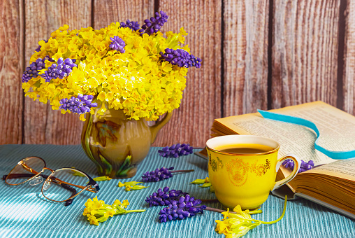 Bouquet of yellow primroses and muscari in a vase. Wooden background, book, yellow cup, glasses. Blue tablecloth.