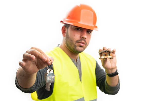 Male builder wearing orange safety hardhat holding wooden miniature house offering home keys close-up selective focus isolated on white background