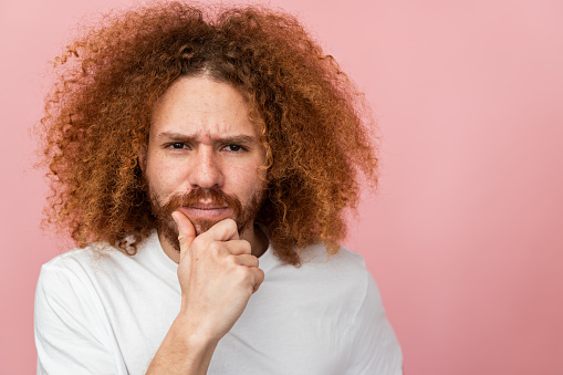 Man with curly hair in thought with hand on chin, contemplating, pink background