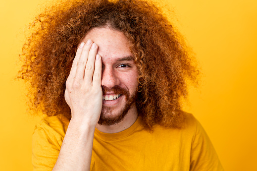 Smiling man with curly hair playfully covering half of face with hand, yellow background