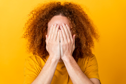 Man with curly hair hiding face with hands, embarrassed or shy, yellow background