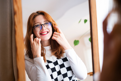 A beautiful woman wearing eyeglasses is looking at herself while sitting in front of the mirror at home.