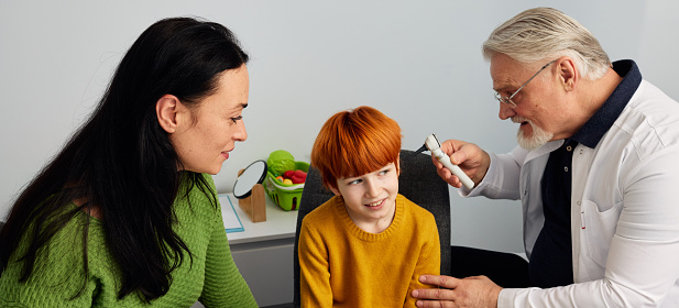 Mother with male child visiting pediatric audiologist for hearing check-up. Red-haired boy having otoscopy in audiology