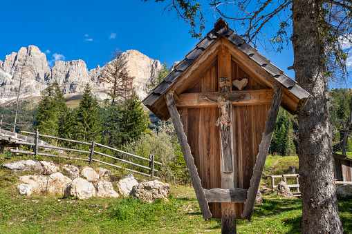 Wooden carving wayside shrine with jesus statue in front of Rosengarten mountain massif, Dolomites, italy. Beautiful blue sky.