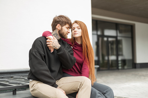 Fashionable beautiful young couple, a handsome hipster man with a beard and tattoo and a lovely redhead stylish woman in a hoodie and pullover are sitting on the street
