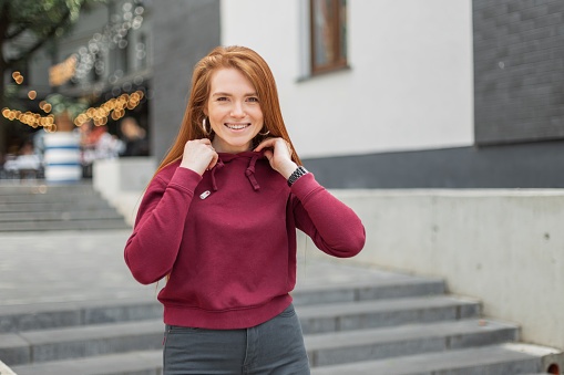 Female cheerful portrait of a beautiful natural fresh fashionable happy redhead girl with a cute smile in a red stylish hoodie walking on the street;. Pretty woman outdoors