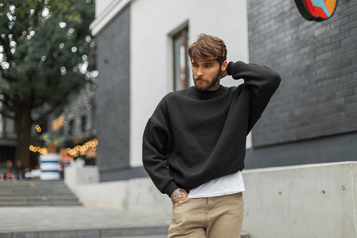 Handsome fashion bearded stylish man in a trendy black sweatshirt mockup walks and straightens his hair on the street