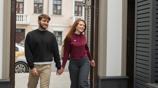 Positive beautiful fashionable couple in fashion urban clothes walks hand in hand in the city. Beautiful happy redhead woman with a smile and a handsome bearded man in a black hoodie walking outdoors