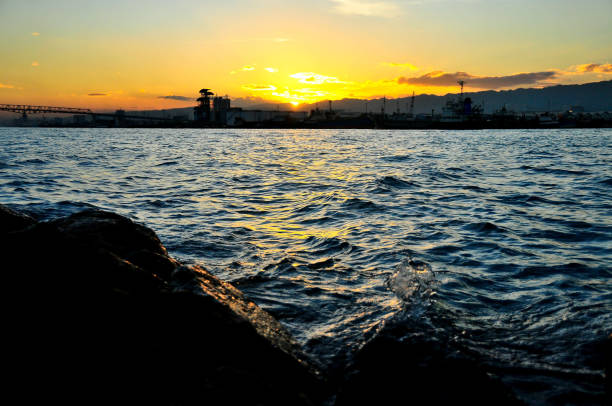 A busy industrial sea port turns into a distant quiet place as the evening twilight unfolds and envelopes the city's skies. The middle-ground sea turns into an oil canvass painting of golden blue color sea ripples against the foreground sea rock.