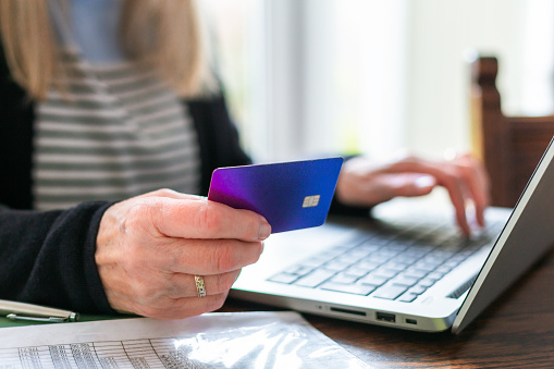 Portrait of a retired senior woman in her 70s making a payment online at home using her smart phone and laptop computer.