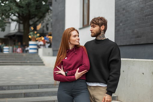 Cool beautiful young fashion couple in fashionable urban clothes with hoodie on the street. Pretty red-haired fresh girl and a handsome brutal bearded man with a tattoo