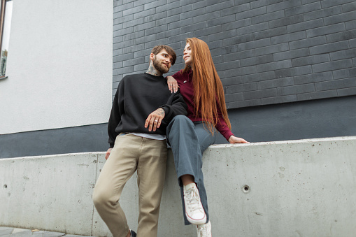Fashionable beautiful stylish couple in fashionable clothes sits and poses near a brick wall on the street. Cool hipster handsome guy with a beard and a pretty red-haired girl with red hoodie