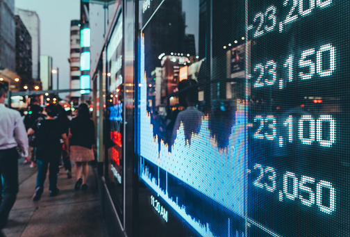 Illuminated stock exchange data on a large outdoor digital display, with blurry people walking by in a city environment during evening hours.