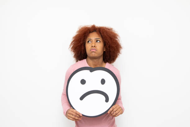 Sadness and disapproval concept - one young woman holding an anthropomorphic sad face. She is looking to the side with an unhappy expression.