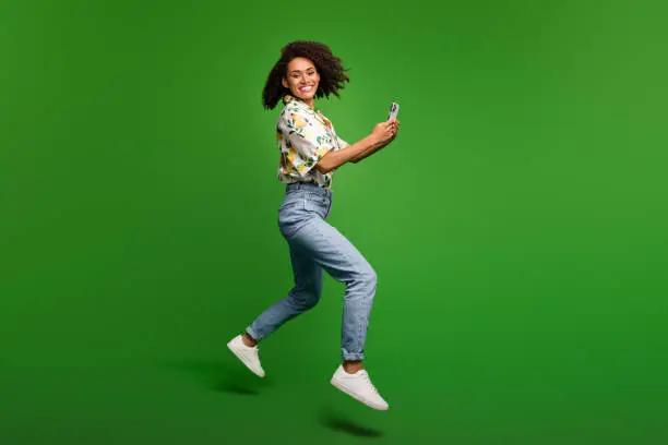 Full length photo of cheerful shiny woman dressed print shirt texting apple iphone samsung gadget jumping isolated green color background.