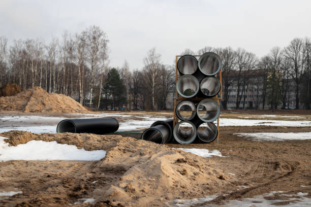 The photograph depicts an industrial or construction scene with large, cylindrical pipes stacked in a pyramid shape. The foreground shows a sandy terrain with tire tracks, and remnants of snow, indicating recent construction activity during a cold season. Bare trees and a building are visible in the background, adding to the feel of an urban construction site in winter.
