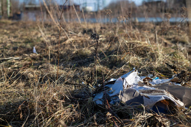 a piece of litter—a cardboard box—amidst other debris on thawing ground during early spring. In the background, industrial structures hint at human activity, while the foreground highlights environmental concerns related to waste and pollution.