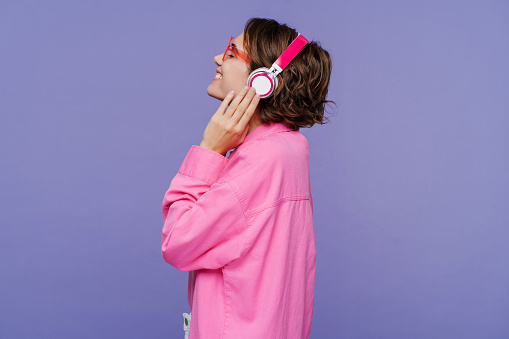 Side view of smiling beautiful woman wearing pink shirt with closed eyes listening to music in headphones, isolated on violet background. Advertisement concept