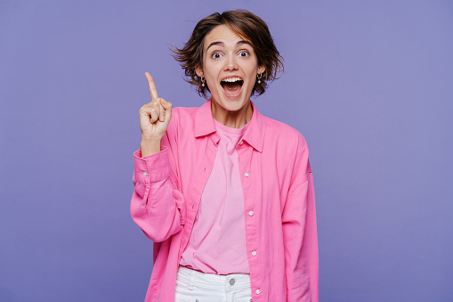 Portrait of smart excited school woman holding finger up, having idea, looking at camera with open mouth isolated on violet background. Education concept