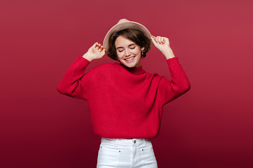 Authentic portrait beautiful smiling young woman wearing stylish comfortable red sweater and hat isolated on red background. Fashion, beauty concept