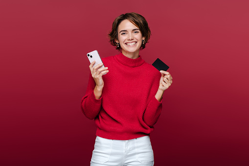 Portrait beautiful smiling woman holding mobile phone, credit card and looking at camera while enjoying shopping online with sales isolated on red background. Internet store, electronic money concept