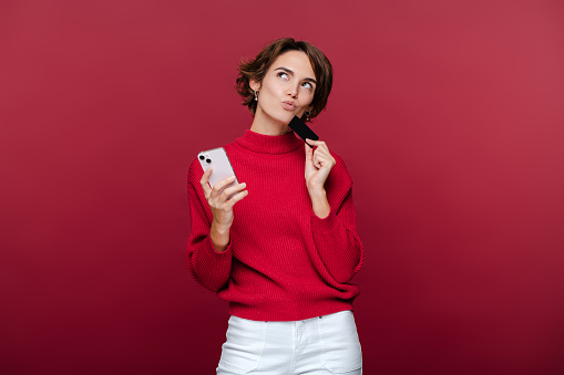 Beautiful pensive woman holding mobile phone and credit card, thinking what to buy, isolated on red background copy space. Concept of mobile banking, technology, online shopping