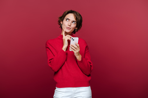 Cute beautiful pensive woman wearing stylish red turtleneck holding mobile phone shopping online looking away isolated on red background. Technology, mobile banking concept