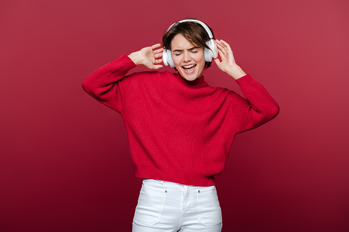 Portrait of attractive young woman wearing stylish casual clothes listening to music in wireless headphones, feeling happy, standing isolated on red background. Technology, positive lifestyle