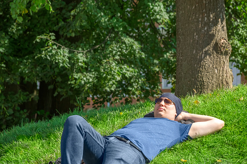 A guy in a bandana and sunglasses lay down to take a nap on a green hill among the greenery