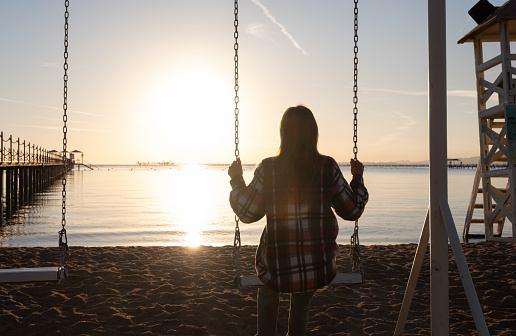 beautiful young woman on the swing on the beach in sunrise and sky background, back view