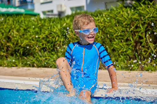 Joyful boy has fun with water sitting on the edge of the pool wearing swimming goggles. Cheerful little kid in a wet bathing suit creates splashes of legs in the pool