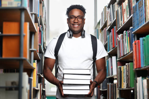 istock Black man stands in the library with a stack of books in his hands and smiles. 2015270854