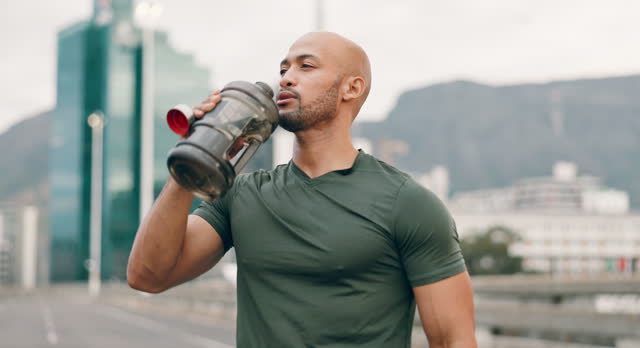 Fitness, running and man drinking water in city for hydration, rest or recovery from cardio workout. Exercise, highway and confident young runner training in urban town with bottle for sports drink