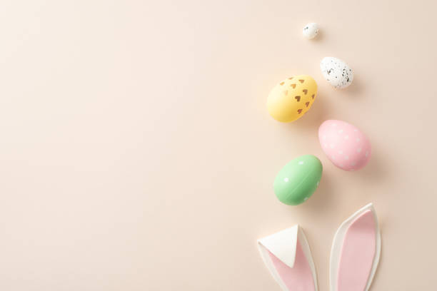istock Festive egg hunt admirer setting. A top view captures lively eggs in flight, accompanied by tucked-away bunny ears on a pastel beige surface, offering space for messages or ads 2015273977