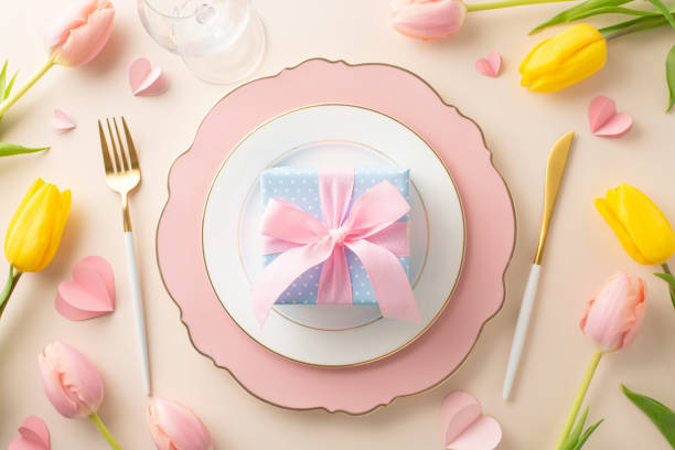 istock Woman's Day banquet setting concept. Top view of refined plate, silverware, wineglass, attractive tulips, gift, and hearts on a light beige base, ideal for advertisement 2015273982