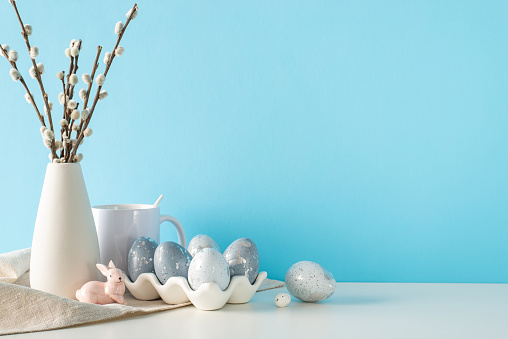 Kitchen table Easter display, side view angle, showcasing a ceramic container with luxurious eggs, a rabbit, mug, and cloth, beside a vase of pussy willow, against a gentle blue wall with text space