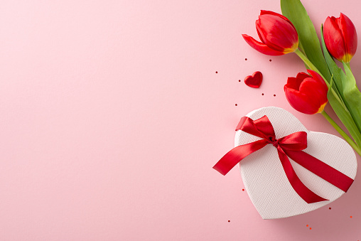 Essential womanly occasion composition. Overhead image of a heart box with a bow, sequins, miniature hearts, and a bunch of red tulips on a gentle pink setting, with space for inscription or publicity