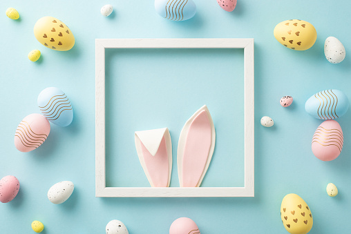 Whimsical Easter magic concept. Top view image featuring an endearing Easter bunny in a photo frame, peeking out with charming ears, alongside vibrant eggs on a baby blue surface