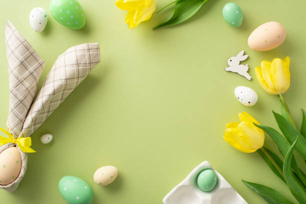 istock Festive Easter display. Top view image showcasing a table arrangement with a bunny-ear wrapped egg, lovely yellow tulips, and egg holder on a soft green background. Perfect for text placement 2015275090