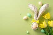 Easter enchantment on display. Top-view photo of table adorned with egg wrapped in bunny ears napkin, vibrant yellow tulips, and sprinkles against soothing pastel green backdrop. Text-friendly layout