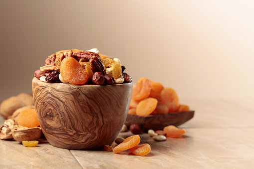 Dried fruits and nuts on a beige ceramic table. The mix of nuts, apricots, and raisins in a wooden bowl.