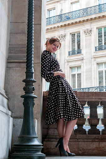 Full length shot of a young brunette in a dotted dress on high heels leaning against wall  in Paris. Medium shot.