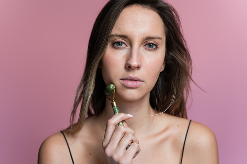 Portrait of a woman massaging her face with a roller on an isolated pink background.