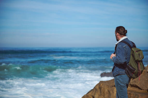 Caucasian young adult bearded hipster man, traveler with backpack, watching the horizon standing on a cliff by the see in a sunny windy day. People. Active lifestyle. Travel. Copy advertising space