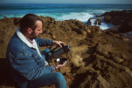 Caucasian bearded young adult man traveler in casual denim, sitting on the rock by sea, enjoying his coffee break in the nature, pouring some hot dink from a stainless steel thermos flag into a mug