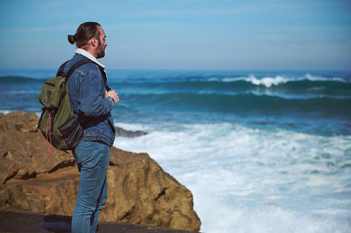 Absorbed man traveling alone, carrying a backpack, looking into the distance, watching horizon, enjoying the view of beautiful nature with splashing waves on the headland, standing alone on the cliff